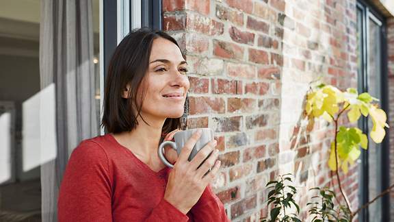 Eine Frau im roten Oberteil steht lächeld vor ihrem Haus in der Sonne und hält einen Kaffeebecher in den Händen