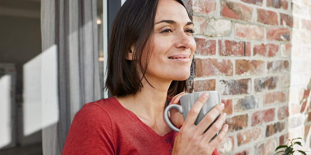 Eine Frau im roten Oberteil steht lächeld vor ihrem Haus in der Sonne und hält einen Kaffeebecher in den Händen