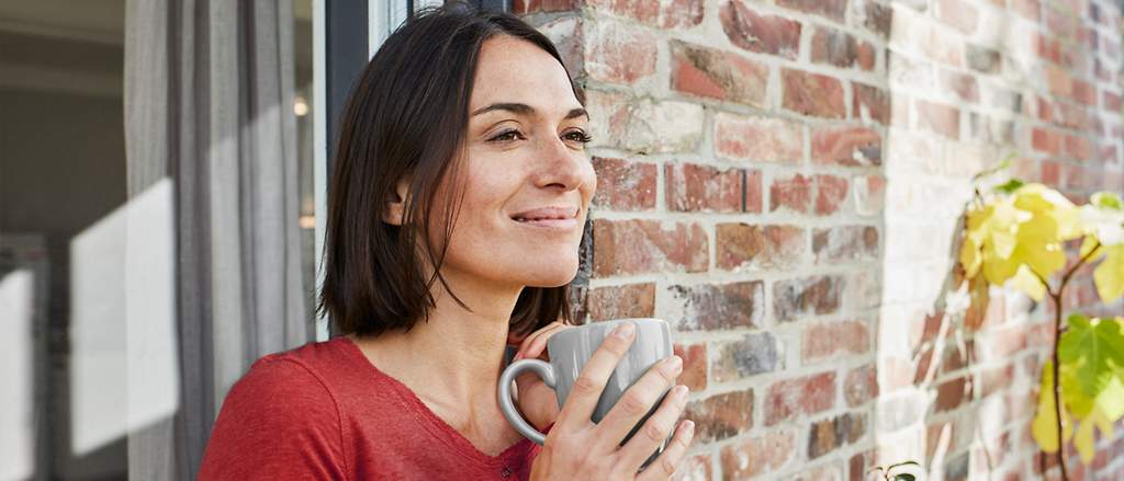 Eine Frau im roten Oberteil steht lächeld vor ihrem Haus in der Sonne und hält einen Kaffeebecher in den Händen