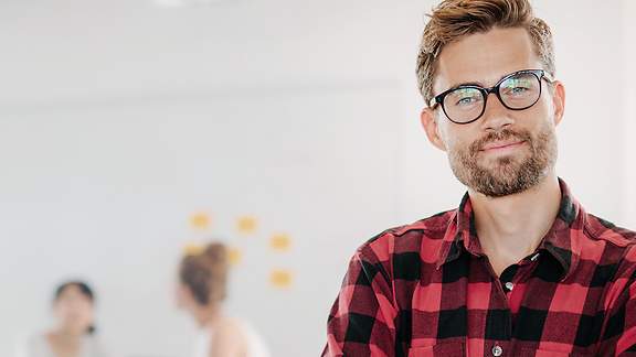 Ein zufriedener junger Mann mit Vollbart und Brille steht mit verschränkten Armen in einem Büro