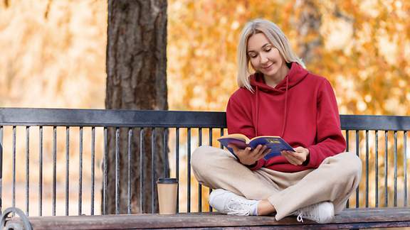 Eine Frau liest auf einer Parkbank unter einem herbstlich verfärbten Baum in einem Buch