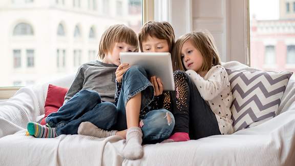 Drei Kinder sitzen auf dem Sofa vor einem Tablet
