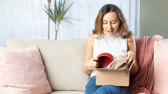 Eine junge Frau im Wohnzimmer öffnet freudig ein Paket, in welchem sich eine rote Schüssel befindet.