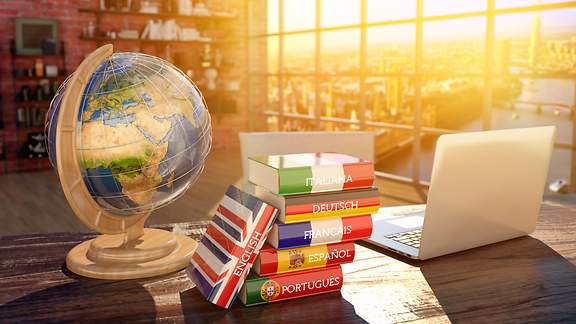 Gestapelte Fremdsprachen-Wörterbücher, ein Laptop und ein Globus stehen auf einem Schreibtisch.