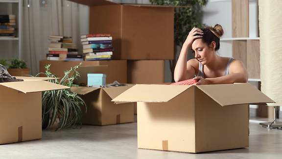 Eine junge Frau sitzt traurig in einer Umzugskiste in einer Wohnung. Im Hintergrund stehen weitere gepackte Umzugskisten.