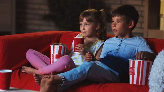 Ein Junge und ein Mädchen sitzen beim perfekten Filmabend mit Popcornbehältern und Getränkebechern auf dem Sofa.