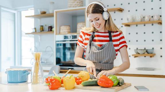 Eine junge Frau mit Kopfhörern schneidet in der Küche fröhlich Gemüse