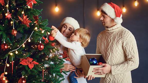 Eine schwangere Frau mit ihrem Sohn auf dem Arm und ein Mann schmücken einen Weihnachtsbaum mit einer Lichterkette.