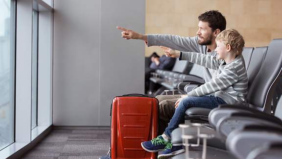 Vater und Sohn sitzen in der Abfluhalle am Flughafen und zeigen auf etwas, das hinter der Scheibe liegt