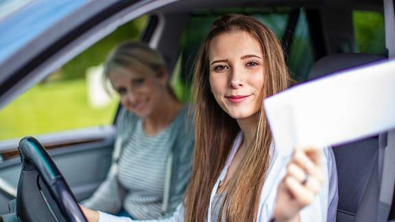 Junge Frau am Steuer eines Wagens mit einer älteren Beifahrerin hält ein Dokument aus dem Fenster