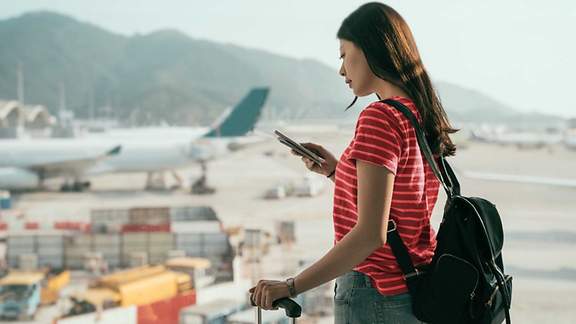 Eine Frau steht mit ihrem Koffer am Flughafen und schaut besorgt in ihr Handy. 