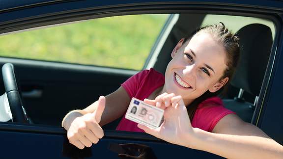 Eine erfreute junge Frau sitzt in einem Auto hält ihren Führerschein in die Kamera