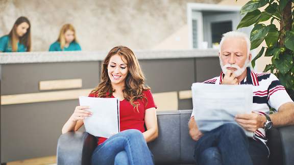 Eine junge Frau und ein älterer Mann lesen im Wartebereich einer Arztpraxis in Unterlagen