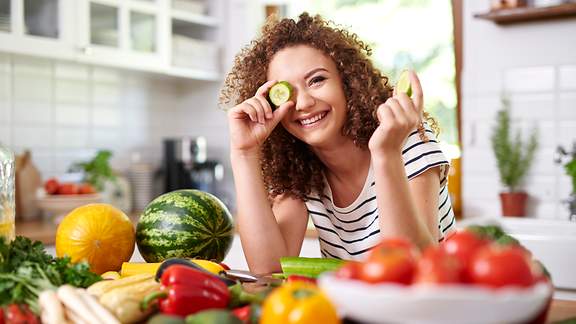 Lachende junge Frau hält sich zwischen Gemüse Gurkenscheibe vors Auge