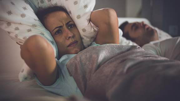 Eine Frau hält sich die Ohren mit ihrem Kopfkissen zu, weil ihr Mann schnarcht.