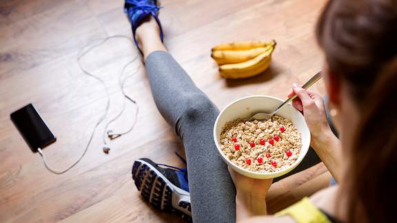 Eine Frau in Sportsachen sitzt auf dem Boden und isst Müsli