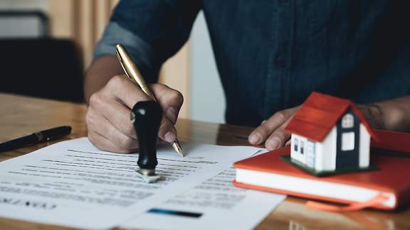 An einem Schreibtisch unterzeichnet ein Mann in dunkelblauem Leinenhemd einen Hypothekenvertrag. 