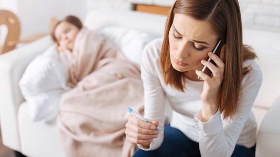 Eine Mutter telefoniert und ihr krankes Kind liegt im Hintergrund auf der Couch.