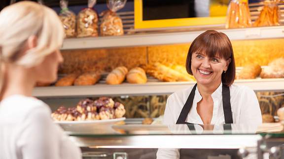 Eine freundlich lächelnde Verkäuferin in einer Bäckerei bedient eine vor der Theke stehende Kundin. 