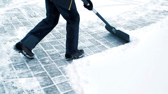 Eine Person befreit Gehwegkacheln mit dem Schneeschieber vom Schnee.