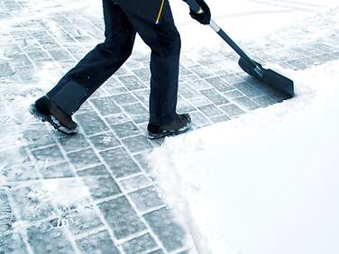 Eine Person befreit Gehwegkacheln mit dem Schneeschieber vom Schnee.