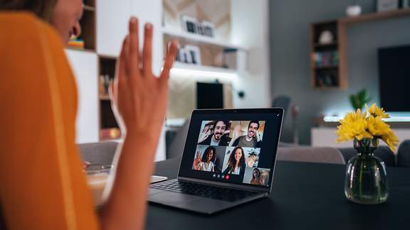 Eine Frau am Laptop winkt ihren Kollegen in der Videokonferenz zu.