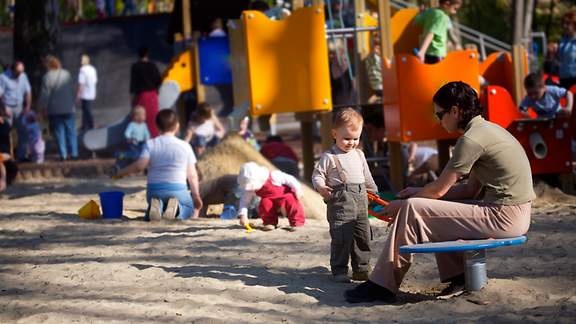 Eine auf einem Spielgerät sitzende Mutter passt auf ihr auf einem Kinderspielplatz spielendes Kleinkind auf.