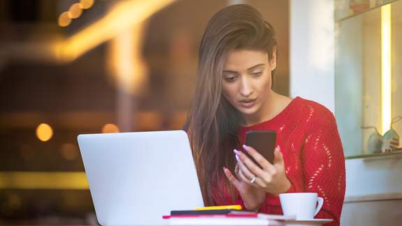 Eine junge Frau sitzt in einem Kaffee und blickt entsetzt auf ihr Smartphone
