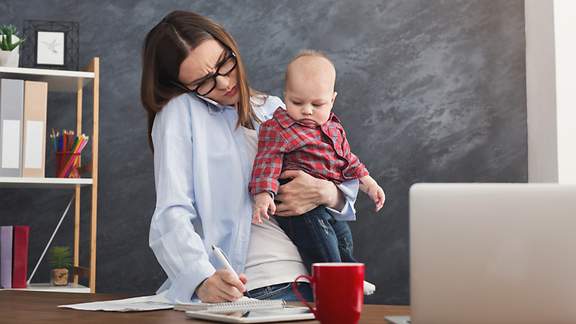 Junge Frau hält ihr Baby in den Armen während sie arbeitet.