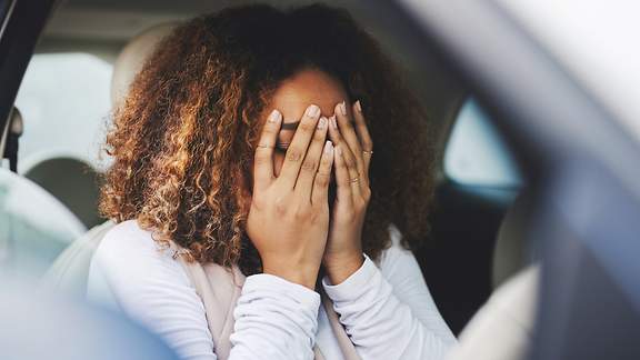 Eine Frau sitzt im Auto und verdeckt ängstlich ihr Gesicht mit beiden Händen.