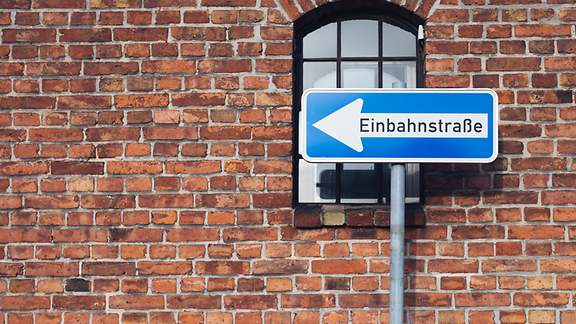 Vor einer Hausmauer mit Fenster steht ein Einbahnstraßenschild.