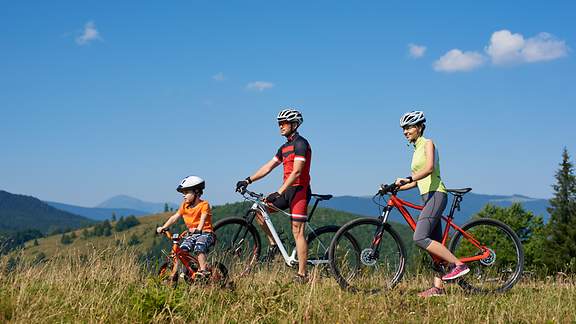 Mutter, Vater und Kind mit Fahrrädern ruhen sich auf dem Gipfel eines grasbewachsenen Hügels aus.