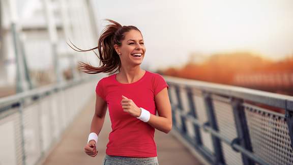 Junge Frau joggt lachend über eine stählerne Fußgängerbrücke