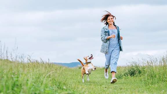 Frau läuft mit ihrem Hund über eine grüne Wiese