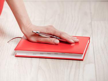 Eine weibliche Hand mit Kugelschreiber liegt auf einem roten Schreibbuch