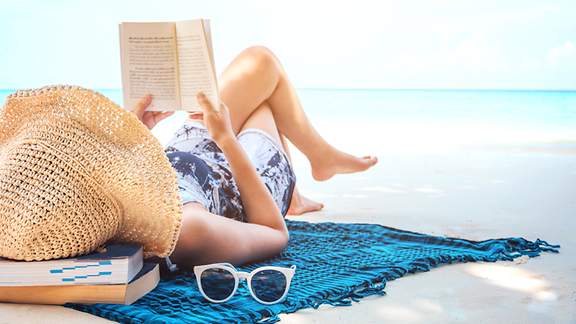 Frau mit Sonnenhut liegt lesend am Strand