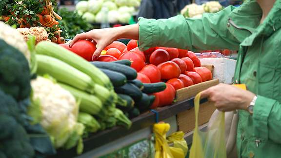 Eine ältere Frau greift auf dem Wochenmarkt nach frischem Gemüse