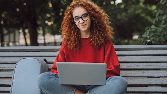 Junge rothaarige Frau mit Brille sitzt mit ihrem Laptop auf dem Schoß auf einer Bank